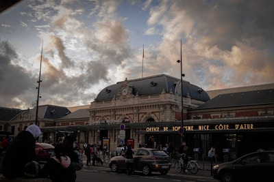 A busy scene outside a large, ornate building with a domed roof and intricate architectural details. The building has signs indicating it is connected to SNCF and the Office de Tourisme Nice Côte d'Azur. Numerous people and vehicles, including bikes and cars, are in front of the building, suggesting a public area or transport hub. The sky above is partly cloudy with a mix of sunlight and shadows, creating a dramatic effect.