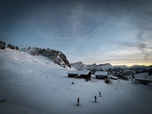 A winter landscape with snow-covered mountains and ski slopes. A few skiers are making their way down the hill toward wooden chalets nestled in the snow. The sky is a gradient of blue, adding tranquility to the scene. Pine trees with snow-dusted branches are visible on the left, and distant peaks can be seen in the background.