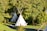Several tipi tents are situated in a lush, green forest environment. A winding gravel path leads through the area, surrounded by dense foliage and trees. The scene is serene and sunlit, with some shadows creating a dappled effect on the grass.
