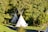 Several tipi tents are situated in a lush, green forest environment. A winding gravel path leads through the area, surrounded by dense foliage and trees. The scene is serene and sunlit, with some shadows creating a dappled effect on the grass.