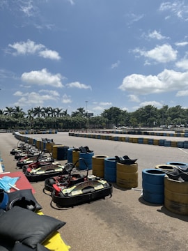 A racetrack is lined with brightly colored barrels and several go-karts are parked in the foreground. The sky is clear with a few clouds, and palm trees are visible in the background, suggesting a warm, sunny day.