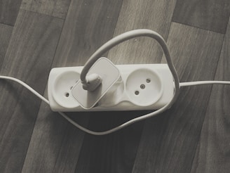 A power strip with three sockets is placed on a wooden floor. One socket is occupied by a white power plug with a cable that loops around. The other two sockets are empty.