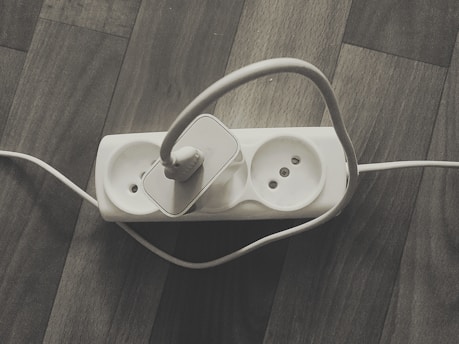 A power strip with three sockets is placed on a wooden floor. One socket is occupied by a white power plug with a cable that loops around. The other two sockets are empty.