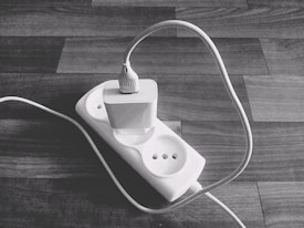 A black and white photo of a power strip placed on a wooden surface with a single plug connected to it. The plug is attached to a long cable that curves and stretches out of the frame.