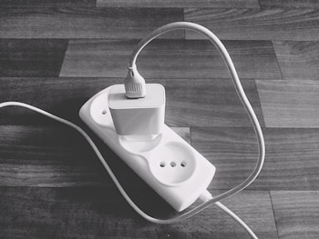 A black and white photo of a power strip placed on a wooden surface with a single plug connected to it. The plug is attached to a long cable that curves and stretches out of the frame.