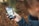 A person is holding a smartphone with a folder labeled 'Kids' open on the screen. The phone shows various apps, some of which are designed for children, including a dinosaur icon and other child-friendly apps. The background is blurred, with autumn leaves on the ground.