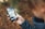 A person is holding a smartphone with a folder labeled 'Kids' open on the screen. The phone shows various apps, some of which are designed for children, including a dinosaur icon and other child-friendly apps. The background is blurred, with autumn leaves on the ground.