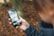 A person is holding a smartphone with a folder labeled 'Kids' open on the screen. The phone shows various apps, some of which are designed for children, including a dinosaur icon and other child-friendly apps. The background is blurred, with autumn leaves on the ground.