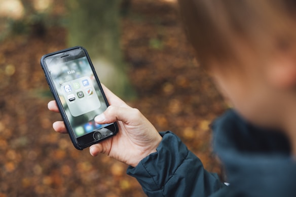 A person is holding a smartphone with a folder labeled 'Kids' open on the screen. The phone shows various apps, some of which are designed for children, including a dinosaur icon and other child-friendly apps. The background is blurred, with autumn leaves on the ground.