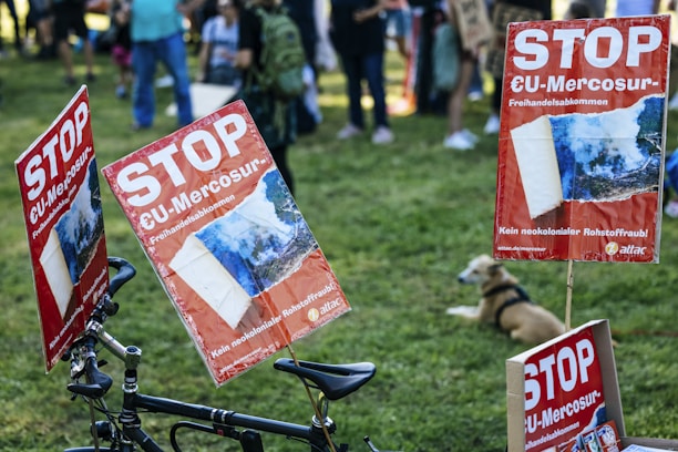 Red protest signs with the text 'STOP EU-Mercosur' are attached to a bicycle. People are gathered in the background on a grassy area, and a dog is lying down nearby. The atmosphere suggests a demonstration or protest gathering.