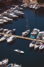 Aerial view of a marina with numerous luxury yachts and boats docked along a pier. The water is a deep blue, and there is a small motorboat moving through the harbor. The dock is adjacent to a promenade lined with shops and buildings.