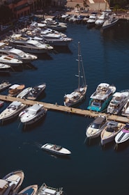 Aerial view of a marina with numerous luxury yachts and boats docked along a pier. The water is a deep blue, and there is a small motorboat moving through the harbor. The dock is adjacent to a promenade lined with shops and buildings.
