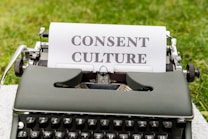 An old-fashioned typewriter with a piece of paper inserted, displaying the words 'Consent Culture', set outdoors on a grassy surface.