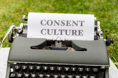 An old-fashioned typewriter with a piece of paper inserted, displaying the words 'Consent Culture', set outdoors on a grassy surface.