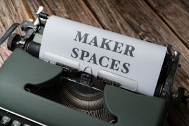 A vintage typewriter with a sheet of paper inserted. The paper has the words 'MAKER SPACES' typed in bold letters. The typewriter is placed on a rustic wooden surface, adding a classic and nostalgic feel.