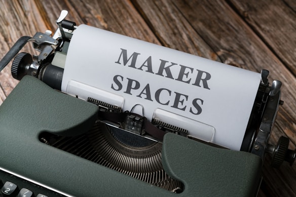 A vintage typewriter with a sheet of paper inserted. The paper has the words 'MAKER SPACES' typed in bold letters. The typewriter is placed on a rustic wooden surface, adding a classic and nostalgic feel.
