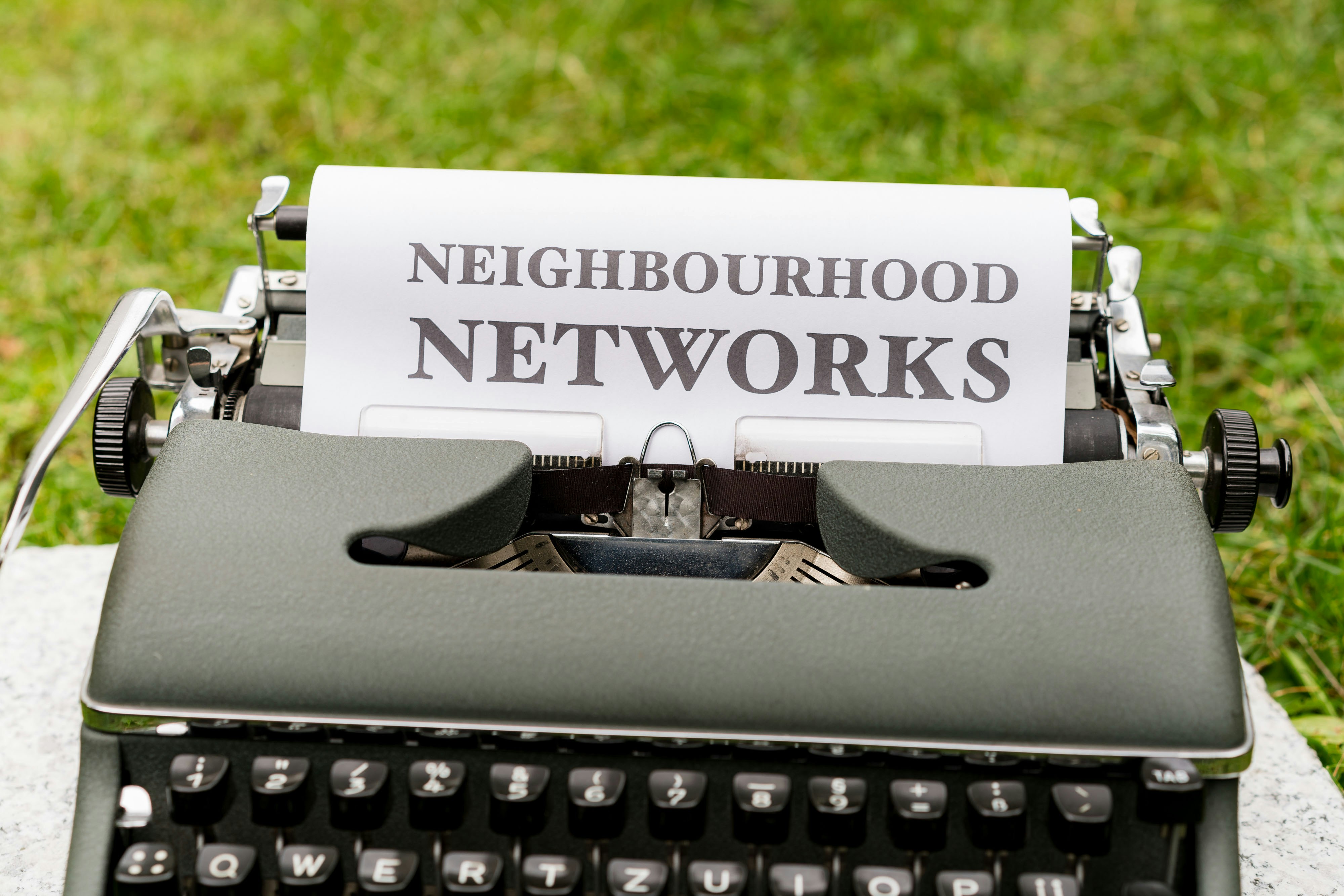 An old-fashioned typewriter is placed on a grassy surface with a sheet of paper inserted into it. The paper features the words 'NEIGHBOURHOOD NETWORKS' typed in bold, uppercase letters.