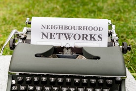 An old-fashioned typewriter is placed on a grassy surface with a sheet of paper inserted into it. The paper features the words 'NEIGHBOURHOOD NETWORKS' typed in bold, uppercase letters.
