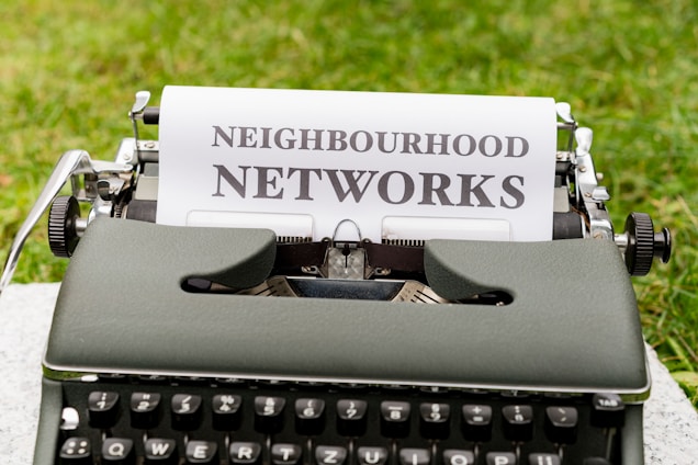 An old-fashioned typewriter is placed on a grassy surface with a sheet of paper inserted into it. The paper features the words 'NEIGHBOURHOOD NETWORKS' typed in bold, uppercase letters.