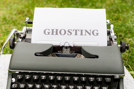 An old-fashioned typewriter holds a sheet of white paper with the word 'GHOSTING' typed in bold, capital letters. The typewriter is set outdoors on a grassy surface, creating a vintage and literary atmosphere.