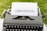 An old-fashioned typewriter holds a sheet of white paper with the word 'GHOSTING' typed in bold, capital letters. The typewriter is set outdoors on a grassy surface, creating a vintage and literary atmosphere.