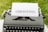 An old-fashioned typewriter holds a sheet of white paper with the word 'GHOSTING' typed in bold, capital letters. The typewriter is set outdoors on a grassy surface, creating a vintage and literary atmosphere.