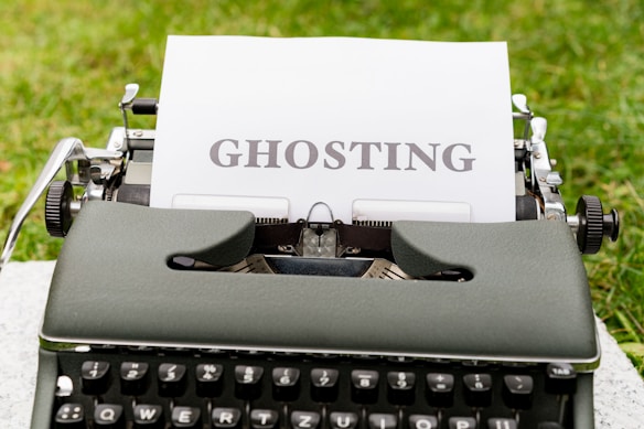An old-fashioned typewriter holds a sheet of white paper with the word 'GHOSTING' typed in bold, capital letters. The typewriter is set outdoors on a grassy surface, creating a vintage and literary atmosphere.