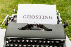 An old-fashioned typewriter holds a sheet of white paper with the word 'GHOSTING' typed in bold, capital letters. The typewriter is set outdoors on a grassy surface, creating a vintage and literary atmosphere.
