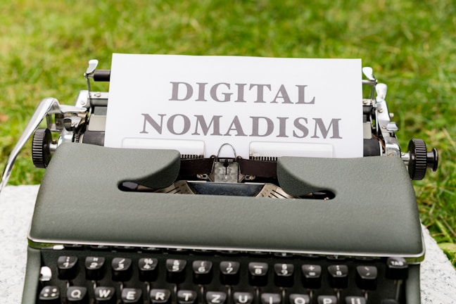 A vintage typewriter set on a grassy surface with a white sheet of paper showing the words 'DIGITAL NOMADISM'. The text is typed in bold, black letters, and the environment suggests an outdoor setting.