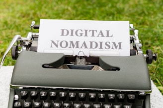 A vintage typewriter set on a grassy surface with a white sheet of paper showing the words 'DIGITAL NOMADISM'. The text is typed in bold, black letters, and the environment suggests an outdoor setting.