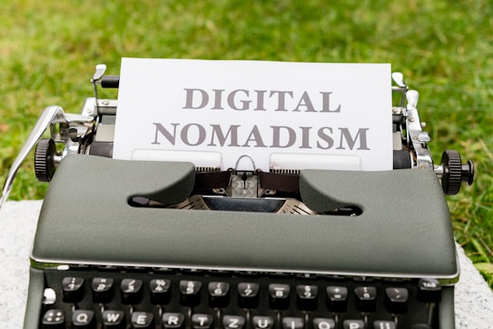 A vintage typewriter set on a grassy surface with a white sheet of paper showing the words 'DIGITAL NOMADISM'. The text is typed in bold, black letters, and the environment suggests an outdoor setting.