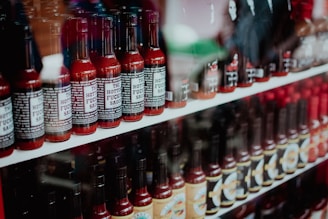 Rows of bottles are displayed on shelves, each filled with a dark red liquid, indicating various types of hot sauces. The bottles have a similar shape and size, with some labels in focus revealing bold text. Reflection on the glass surface creates a slightly distorted view of the products.