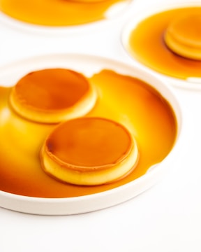 A close-up view of a dessert, likely flan or crème caramel, with a smooth caramel layer on top. The dessert is presented on a white plate with caramel sauce surrounding it. The surface is glossy and the caramel is a rich golden brown.
