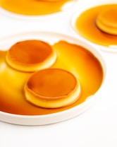 A close-up view of a dessert, likely flan or cr&egrave;me caramel, with a smooth caramel layer on top. The dessert is presented on a white plate with caramel sauce surrounding it. The surface is glossy and the caramel is a rich golden brown.