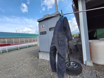 A dark blue or black coverall hangs on a clothesline by a blue clip, next to a metal utility box and a spare tire. The background includes a farm building with a green roof, white fence, and a clear blue sky with clouds.