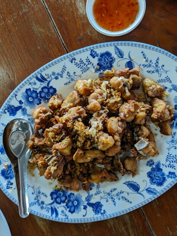 A plate of fried chicken pieces garnished with crispy garlic and placed on a white plate with blue floral patterns. A metal spoon rests on the side of the plate. A small bowl filled with orange dipping sauce is placed beside the plate, set on a wooden table.