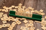 Several wooden letter tiles are scattered across a wooden surface, with one set of tiles arranged on a green tile holder to form the word 'FIGHT'. The tiles show various letters and point values.