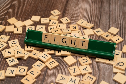Several wooden letter tiles are scattered across a wooden surface, with one set of tiles arranged on a green tile holder to form the word 'FIGHT'. The tiles show various letters and point values.