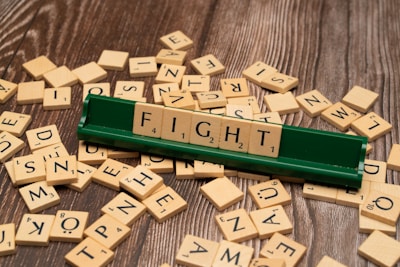 Several wooden letter tiles are scattered across a wooden surface, with one set of tiles arranged on a green tile holder to form the word 'FIGHT'. The tiles show various letters and point values.