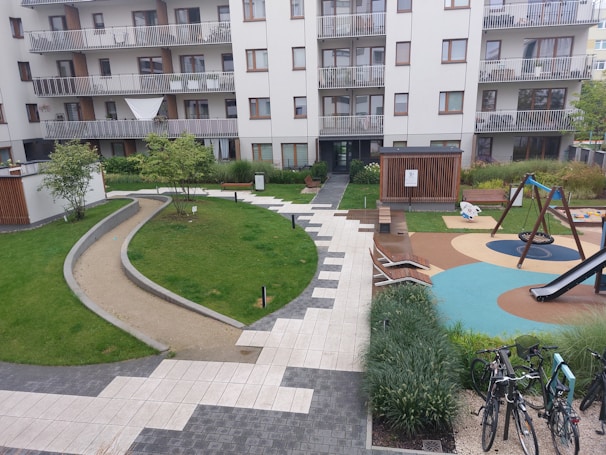 A modern apartment complex with multiple balconies. The courtyard features a playground with swings and a slide, surrounded by greenery and pathways. Wooden benches and lounge chairs are placed around the space. Several bicycles are parked in racks nearby.
