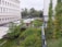 A lush green rooftop garden with various shrubs and grasses is visible. The garden features benches and is surrounded by a white building with multiple windows. A white car is parked on the pavement nearby. Trees and other vegetation line the background, giving a sense of an urban environment with greenery.