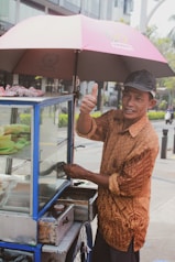 A street vendor is giving a thumbs-up while standing beside his food cart, which is shaded by a red and black umbrella. The vendor wears a patterned brown shirt and a black cap, and he has a friendly expression. The food cart has a glass display with various items inside, and it is mounted on a wheel for mobility. The background shows a modern urban setting with some greenery and a few people in the distance.
