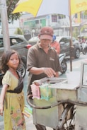 A street vendor operates a cart under a large umbrella on a city street. The vendor, wearing a cap and brown shirt, interacts with a young girl who is holding several snacks. Cars and motorbikes are visible in the background, along with a fence and trees lining the urban setting.