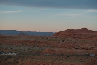 A vast desert landscape with reddish-brown rock formations and sparse vegetation under a clear, pale sky. In the foreground, there are vehicles parked with faint road markings.