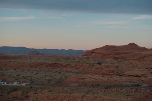 A vast desert landscape with reddish-brown rock formations and sparse vegetation under a clear, pale sky. In the foreground, there are vehicles parked with faint road markings.
