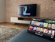 A modern living room features a flat-screen television mounted on a light-colored wooden stand with a patterned wall backdrop. Below the TV, a carpet lies on a wooden floor. In the foreground, a tablet displays a streaming platform interface with various show thumbnails, suggesting a relaxed entertainment setup.