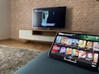 A modern living room features a flat-screen television mounted on a light-colored wooden stand with a patterned wall backdrop. Below the TV, a carpet lies on a wooden floor. In the foreground, a tablet displays a streaming platform interface with various show thumbnails, suggesting a relaxed entertainment setup.