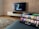 A modern living room features a flat-screen television mounted on a light-colored wooden stand with a patterned wall backdrop. Below the TV, a carpet lies on a wooden floor. In the foreground, a tablet displays a streaming platform interface with various show thumbnails, suggesting a relaxed entertainment setup.