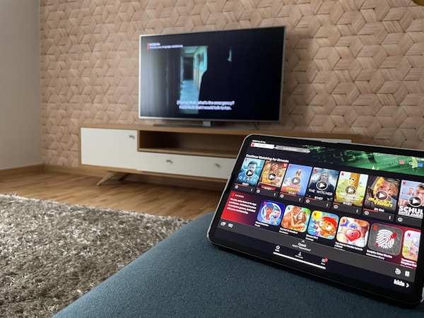 A modern living room features a flat-screen television mounted on a light-colored wooden stand with a patterned wall backdrop. Below the TV, a carpet lies on a wooden floor. In the foreground, a tablet displays a streaming platform interface with various show thumbnails, suggesting a relaxed entertainment setup.