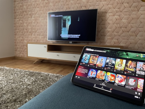 A modern living room features a flat-screen television mounted on a light-colored wooden stand with a patterned wall backdrop. Below the TV, a carpet lies on a wooden floor. In the foreground, a tablet displays a streaming platform interface with various show thumbnails, suggesting a relaxed entertainment setup.
